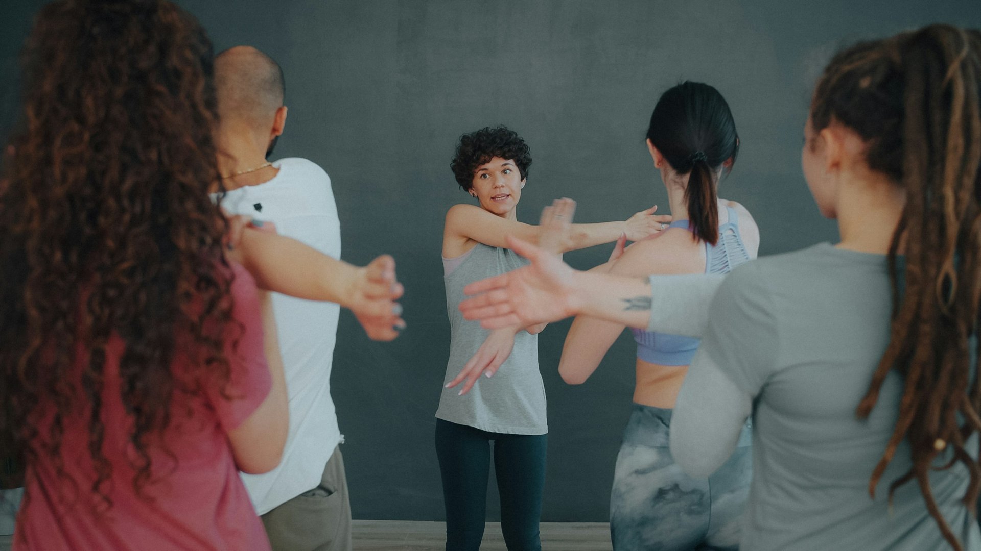 Dans un studio au mur gris, une coach aux cheveux courts et bouclés, face à son groupe, montre un étirement des bras à quatre élèves vus de dos.