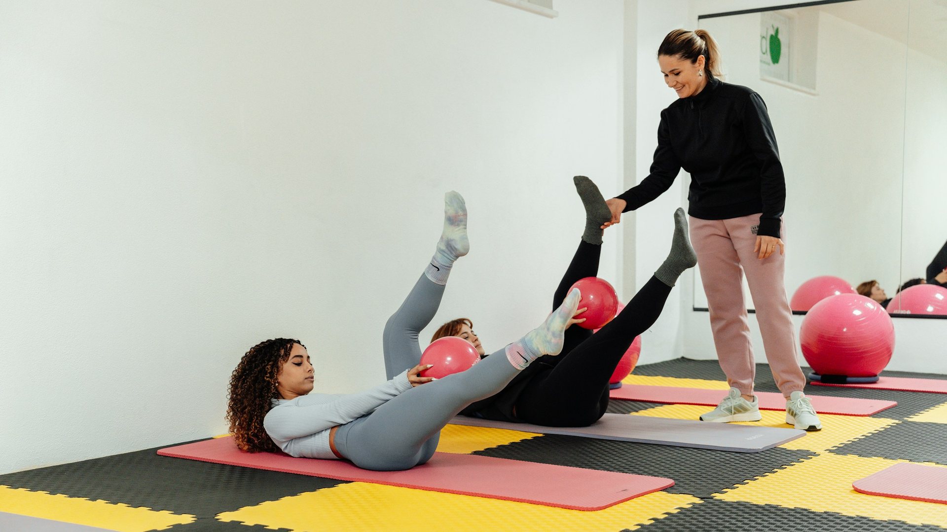 Dans un studio clair, une enseignante debout corrige la jambe levée d'une élève allongée sur un tapis ; une seconde élève, au premier plan, tient un petit ballon rouge.
