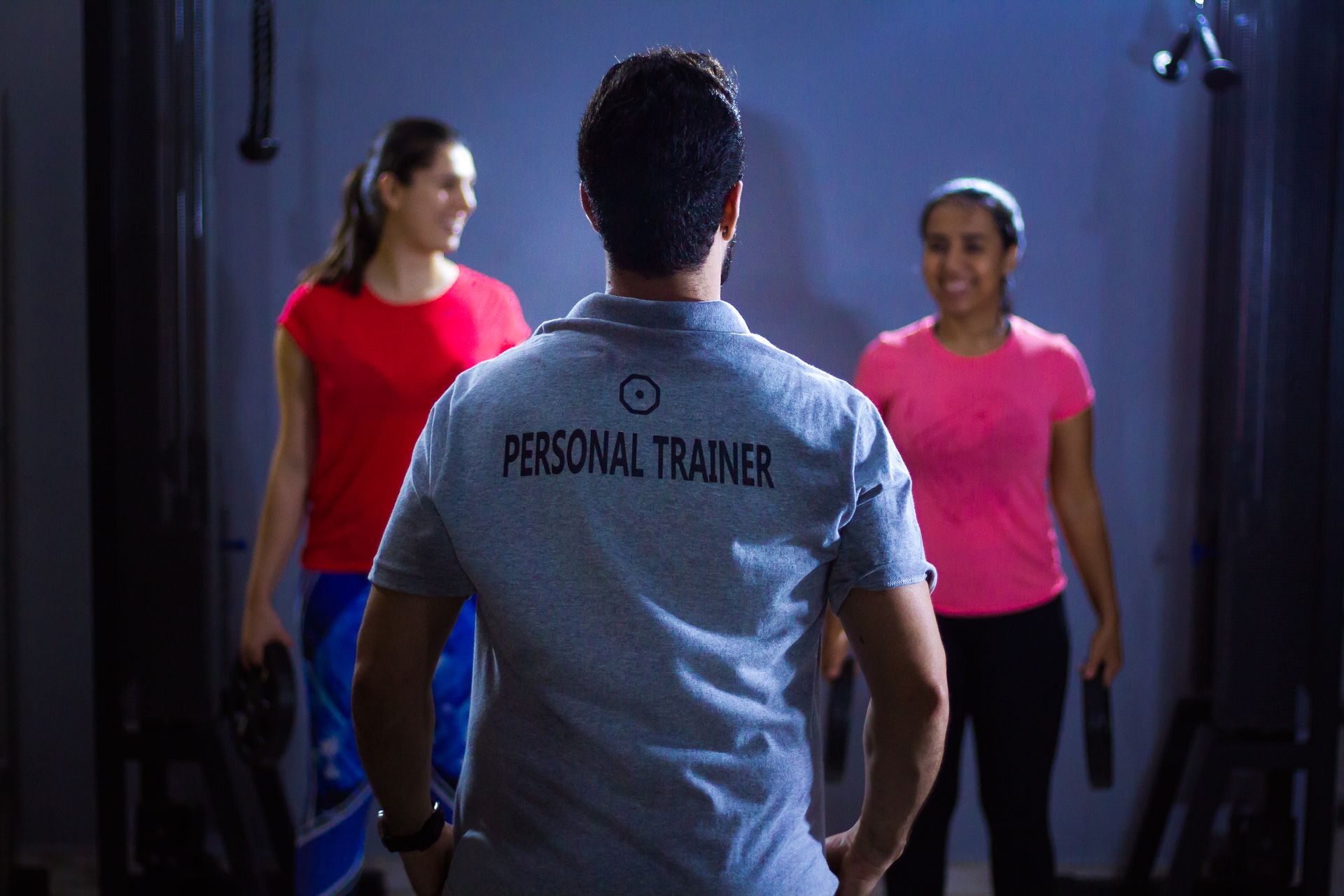Un coach sportif en polo Personal Trainer encadre deux clientes haltères en main dans une salle de sport