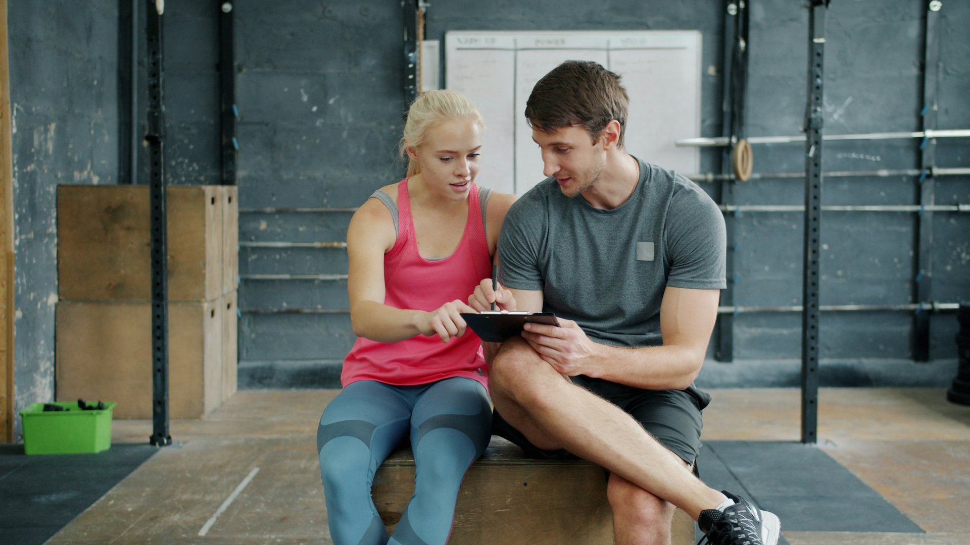 Dans une salle de sport, un coach assis à côté d'une pratiquante note son programme sur un porte-bloc qu'elle montre du doigt.
