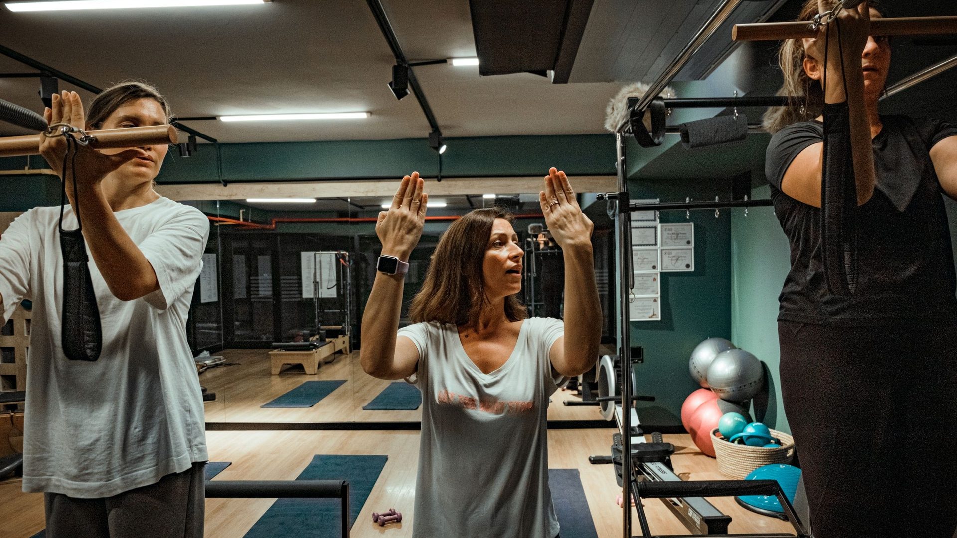 Dans un studio de Pilates aux murs verts, une instructrice montre un mouvement des bras à deux clientes qui tiennent la barre d'un appareil.
